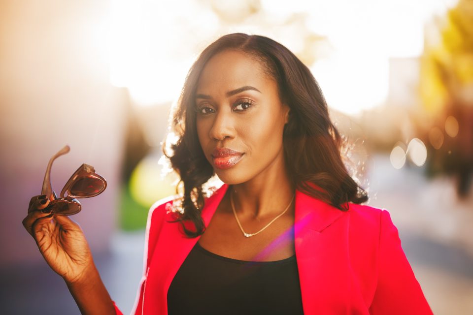 Woman in a bright pink blazer holds sunglasses, standing outdoors with sunlight illuminating her face and a blurred background.