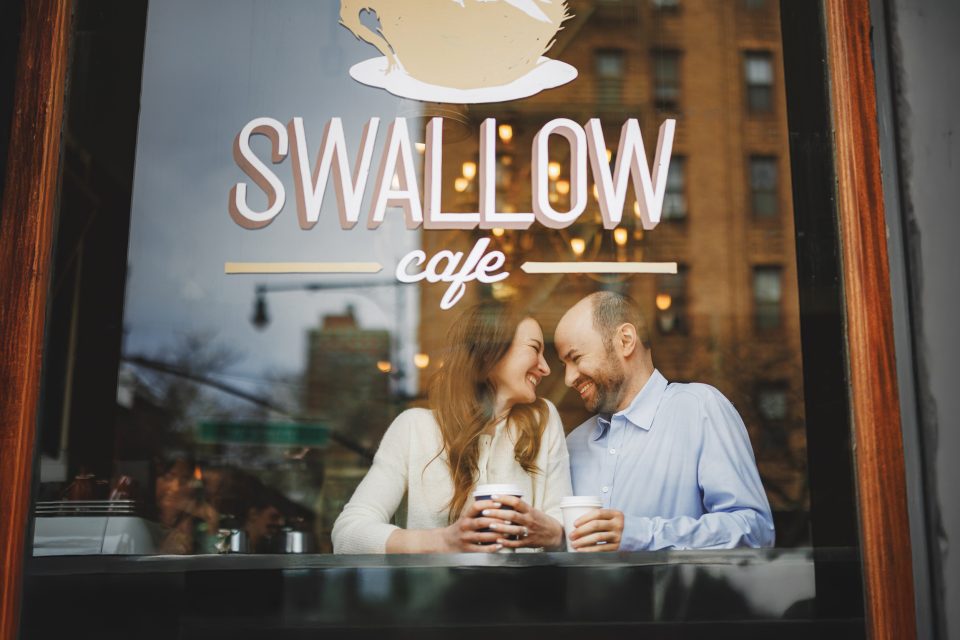 A man and woman sit together indoors, smiling and holding coffee cups at a window counter in Swallow Cafe.