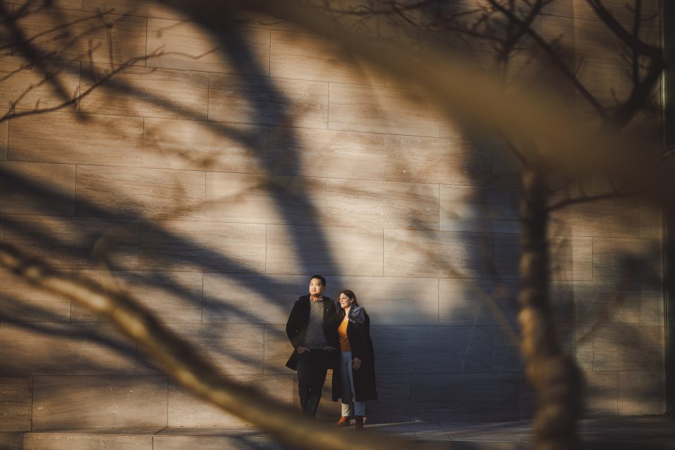Two people stand against a sunlit stone wall, with tree branches and their shadows in the foreground.