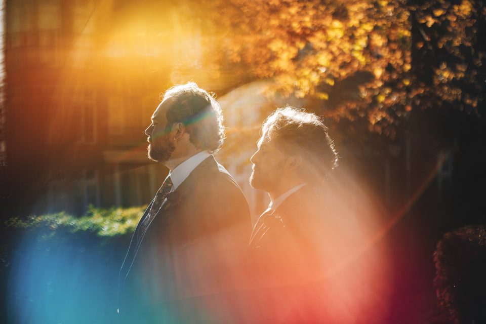 Two men in suits stand outdoors in sunlight, surrounded by colorful light flares and autumn foliage in the background.
