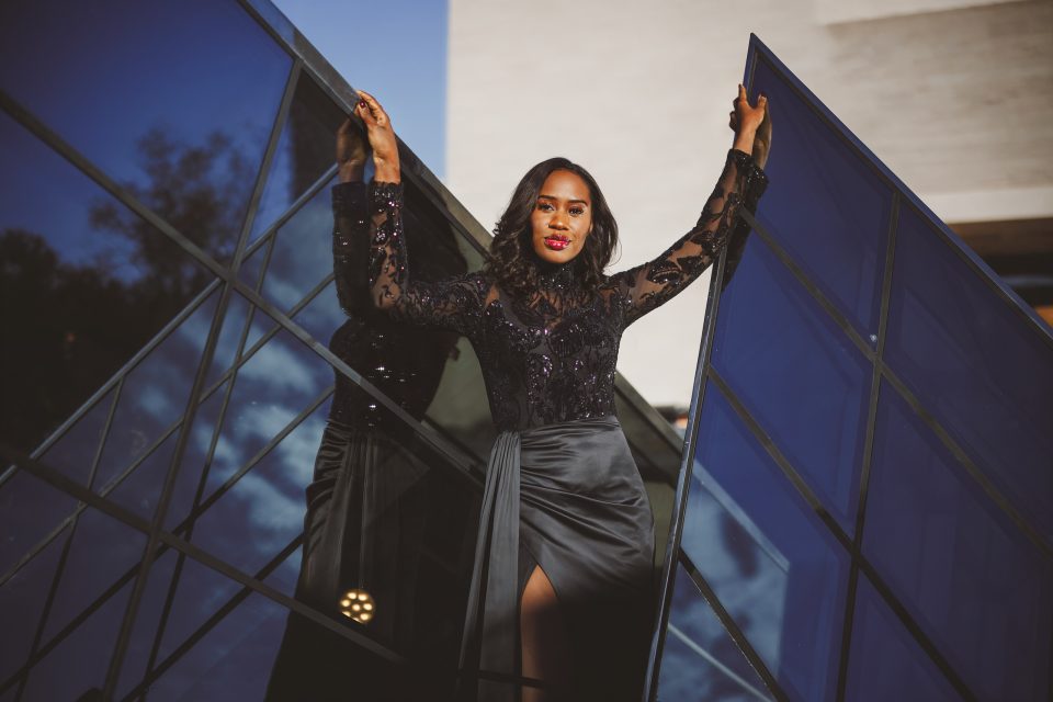A woman in a black dress with sequin details poses with her arms raised, standing between two large glass panels outdoors.