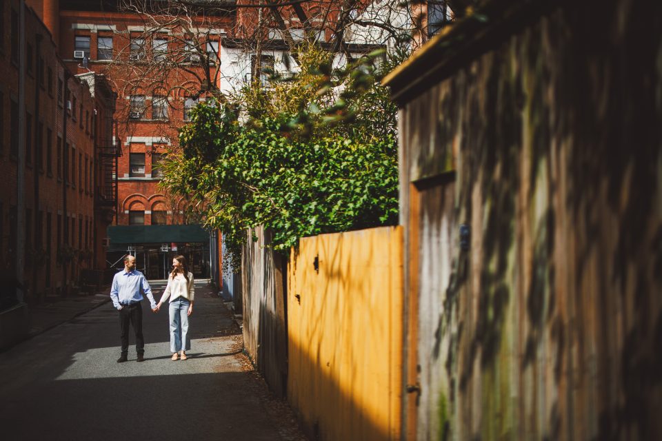 Two people walk hand in hand down a narrow urban alleyway lined with buildings and wooden fences, with sunlight casting shadows.
