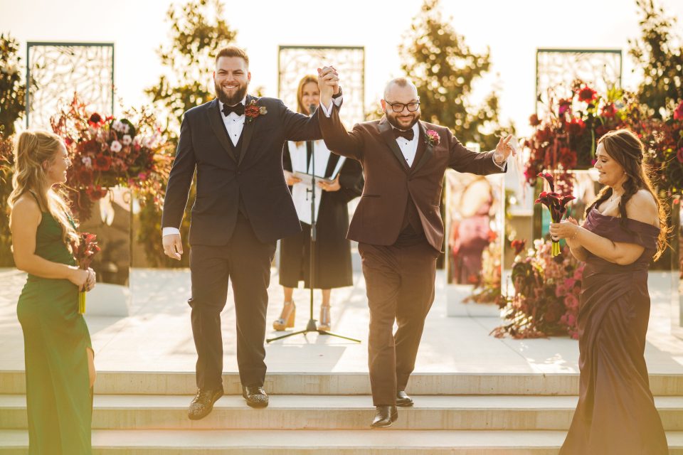 Two men in suits hold hands and smile as they walk down steps at an outdoor wedding ceremony, with two women in dresses nearby and floral arrangements in the background.